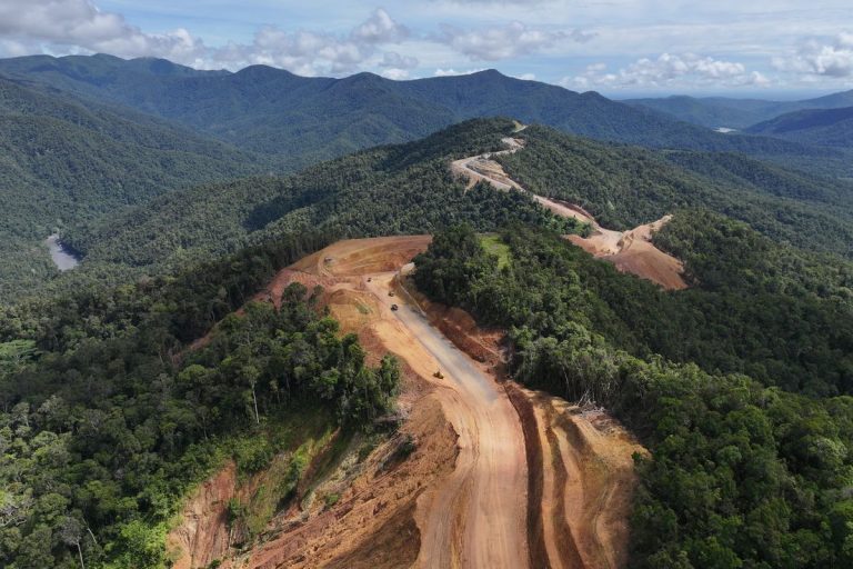 Aerial drone view of a unpaved dirt road winding through dense tropical rainforest mountains in Papua for the Trans-Papua Highway project.
