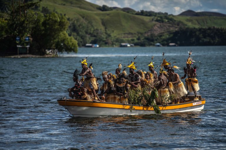People in traditional dress and feather headdresses riding a motorboat on a lake
