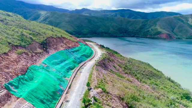 A road along a lake, with green netting holding the hillside above it