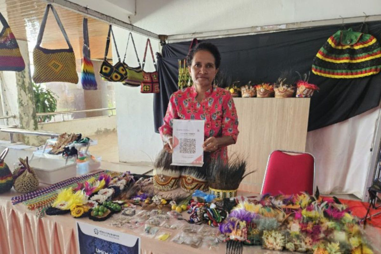 Woman at a market stall holding up a payment QR code beside handicrafts.