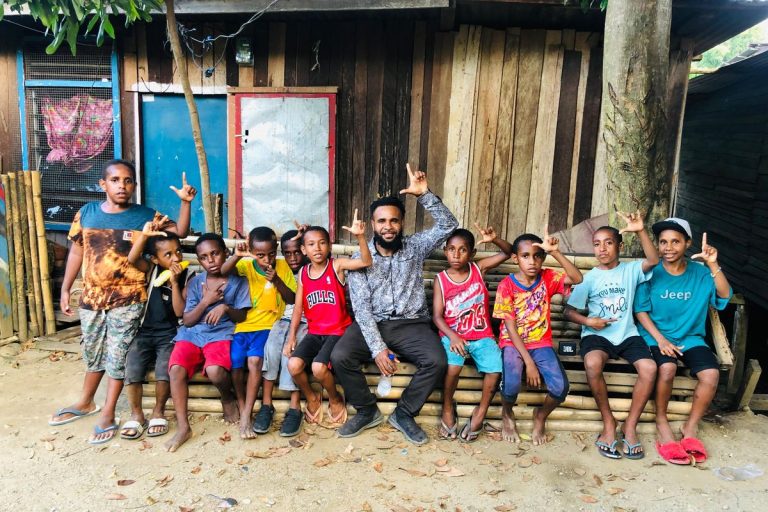 Children and a young man sitting on a bench in front of wooden houses.