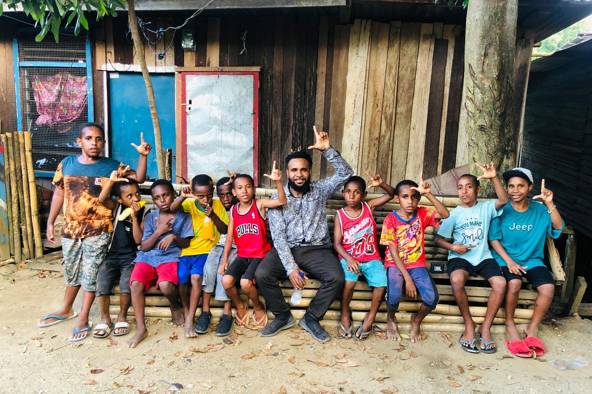 Children and a young man sitting on a bench in front of wooden houses.