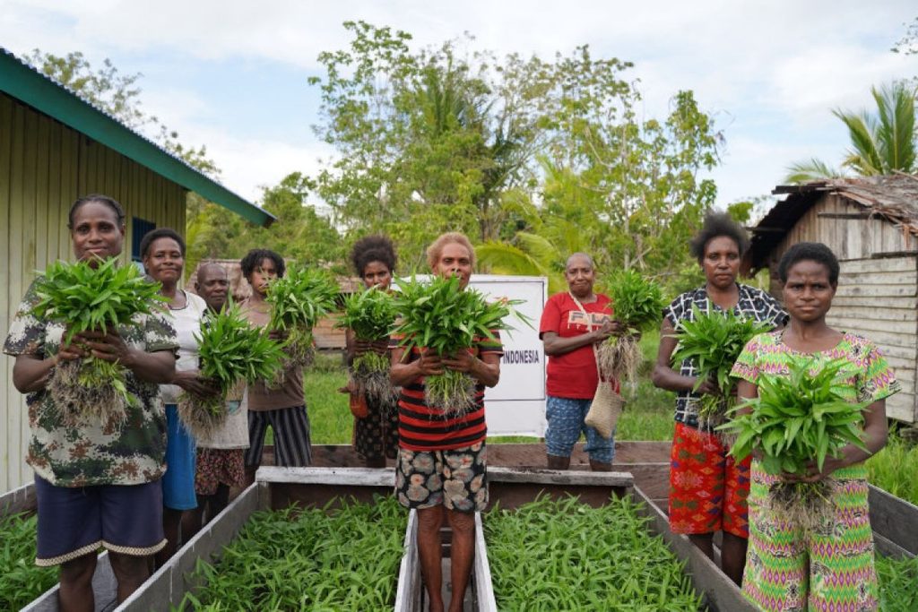 Women holding freshly harvested leafy greens beside raised garden beds.