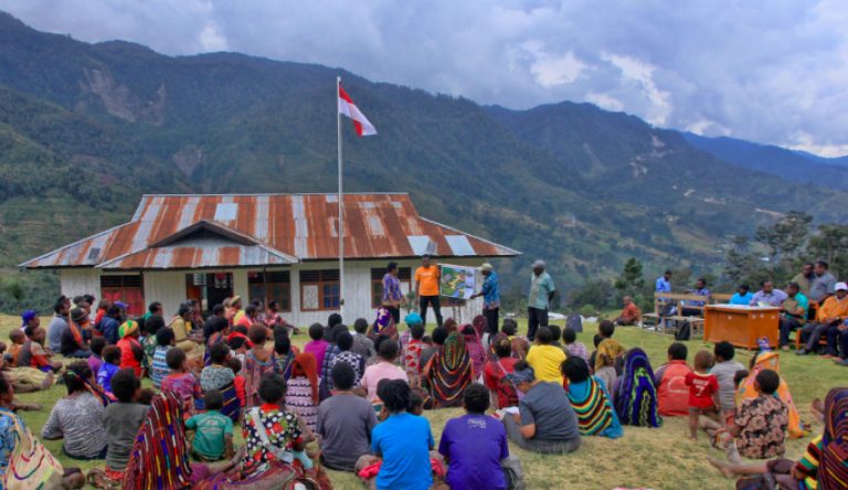 Community gathering on a field in front of a building with an Indonesian flag, mountains behind.