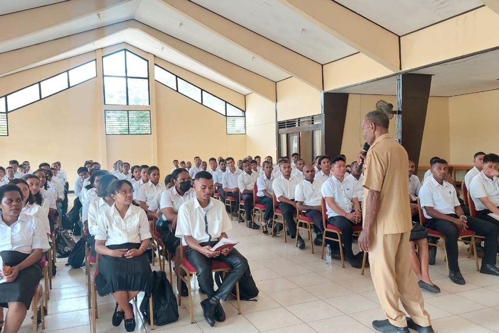 Trainees in white shirts listening to an instructor in a hall.