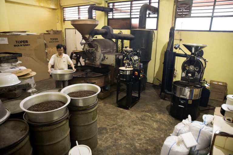 Worker operating coffee roasting machines in a small processing room.