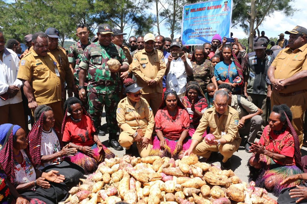 Officials and villagers seated around a pile of sweet potatoes at an event.