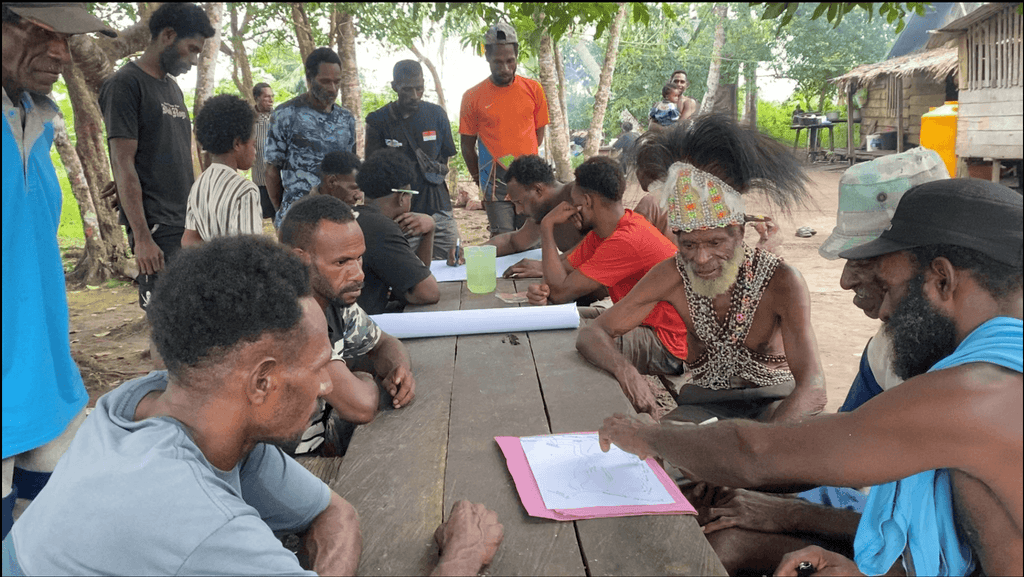 Villagers seated around a wooden table reviewing papers at an outdoor meeting